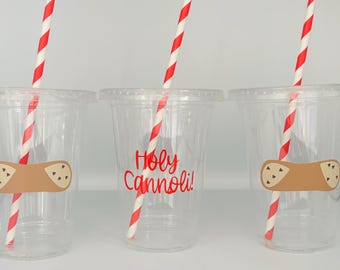 Può includere: Tre bicchieri di plastica trasparente con cannucce a righe rosse e bianche. Ogni bicchiere ha un adesivo a forma di cannolo marrone. La tazza centrale ha la scritta "Holy Cannoli!" in rosso. I bicchieri sono su una superficie bianca.