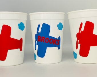 May include: Three white plastic cups with airplane designs. One cup has a red airplane, one has a blue airplane with the name "BROOKS" on it, and the other has a red airplane.
