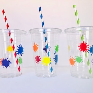 May include: Three clear plastic cups with colorful paint splatter designs and striped straws. The straws are red and white, blue and white, and green and white.