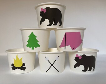 May include: Six white paper cups with different camping-themed designs. The designs include a bear with a pink bow, a pine tree, a tent, a campfire, and two crossed sticks.