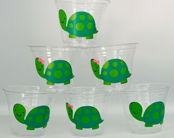 May include: Six clear plastic cups with green turtle designs. Each turtle has a pink bow and a smiling face. The turtles are all facing the same direction.