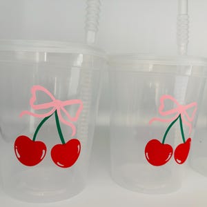 May include: Two clear plastic cups with lids and straws. Each cup has a red cherry with a green stem and a pink bow on top.