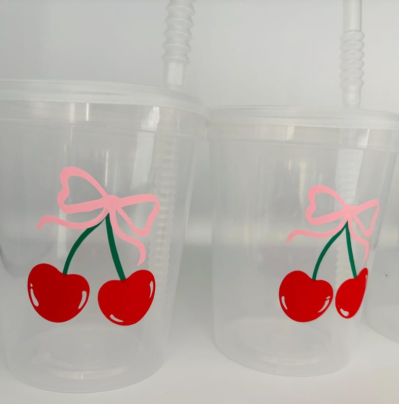 May include: Two clear plastic cups with lids and straws. Each cup has a red cherry with a green stem and a pink bow on top.