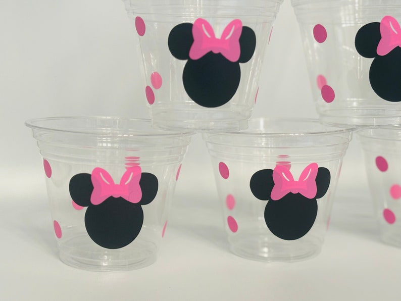 Puede incluir: Cuatro vasos de pl&aacute;stico transparentes con lunares rosas y un dise&ntilde;o de Minnie Mouse negro y rosa.