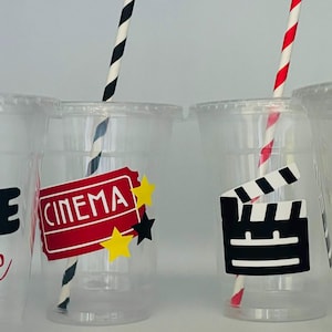 May include: Four clear plastic cups with black and white striped straws. Each cup has a different movie-themed design: "Movie Time" with a film reel, "Cinema" with a red ticket, a clapperboard, and a popcorn bucket.