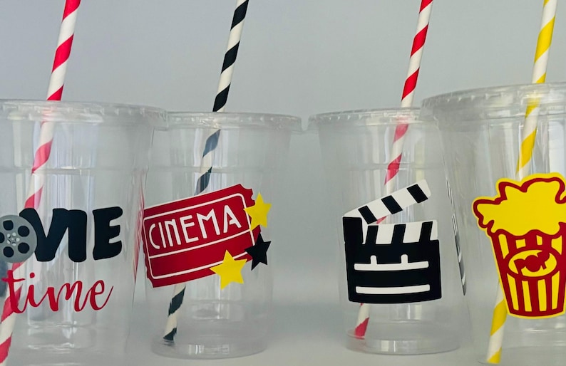 May include: Four clear plastic cups with black and white striped straws. Each cup has a different movie-themed design: "Movie Time" with a film reel, "Cinema" with a red ticket, a clapperboard, and a popcorn bucket.