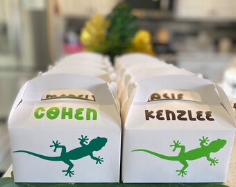 May include: Two white favor boxes with green lizard silhouettes and the names "Cohen" and "Kenzlee" printed in brown letters. The boxes are sitting on a green leaf background.