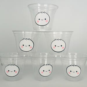 May include: Six clear plastic cups with a cute cartoon dumpling design. Each cup has a white dumpling with black eyes and a smiling mouth, and pink cheeks.