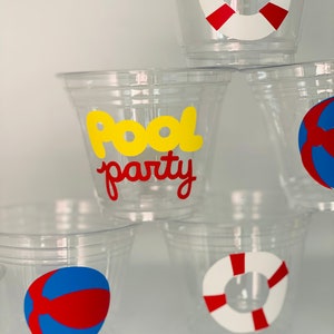 May include: Four clear plastic cups with different pool party themed designs. One cup has a red and blue beach ball, one has a red and white life preserver, one has a red and white life preserver, and one has the words "Pool Party" in yellow letters.