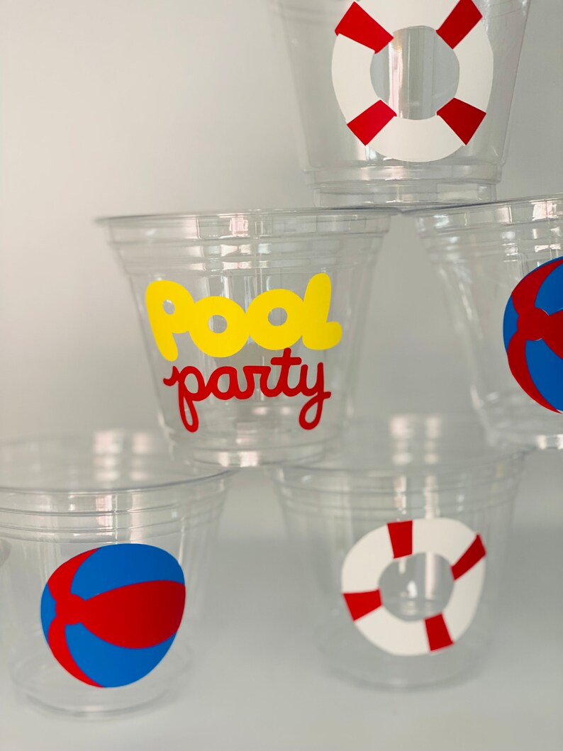 May include: Four clear plastic cups with different pool party themed designs. One cup has a red and blue beach ball, one has a red and white life preserver, one has a red and white life preserver, and one has the words "Pool Party" in yellow letters.
