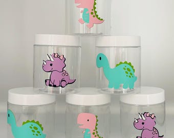 May include: Clear plastic jars with white lids, each decorated with a colorful dinosaur design. The dinosaurs are pink, teal, and purple, with green accents. The jars are arranged in a stack.
