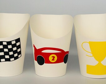 May include: Three off-white paper cups with racing-themed designs. One cup features a black and white checkered flag, another a red race car with the number 2, and the third a yellow trophy. These cups are ideal for a racing-themed party.