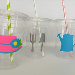 Pu&ograve; includere: Tre bicchieri di plastica trasparente con cannucce colorate e adesivi a tema. Il primo bicchiere ha un cappello rosa con un fiore blu, il secondo bicchiere ha una forchetta e una pala grigie, e il terzo bicchiere ha un annaffiatoio blu.
