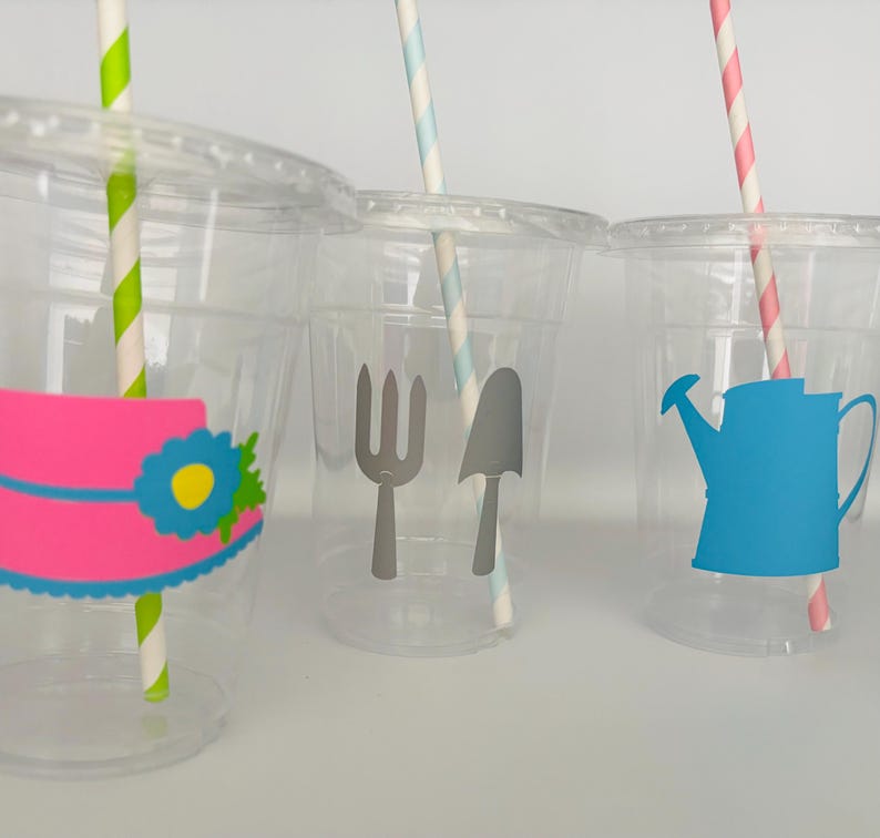 Pu&ograve; includere: Tre bicchieri di plastica trasparente con cannucce colorate e adesivi a tema. Il primo bicchiere ha un cappello rosa con un fiore blu, il secondo bicchiere ha una forchetta e una pala grigie, e il terzo bicchiere ha un annaffiatoio blu.