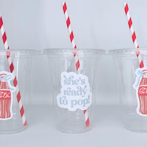 May include: Three clear plastic cups with red and white striped straws. Each cup has a sticker of a red Coca-Cola bottle with a blue bow. The middle cup has the text "she's ready to pop!"