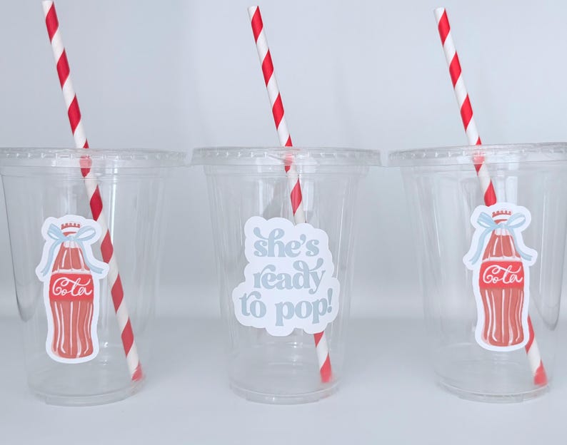 May include: Three clear plastic cups with red and white striped straws. Each cup has a sticker of a red Coca-Cola bottle with a blue bow. The middle cup has the text "she's ready to pop!"