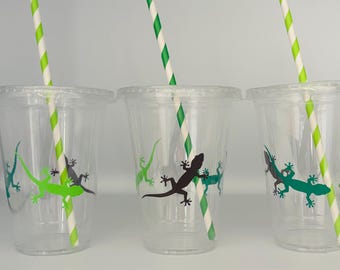 May include: Three clear plastic cups with lids and green and white striped straws. Each cup has a different colored lizard graphic. The lizards are green and black. The straws are green and white.