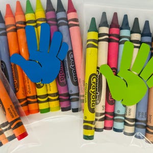 May include: Two clear plastic bags filled with Crayola crayons. The bag on the left has a blue handprint sticker and the bag on the right has a green handprint sticker. The crayons are arranged in a rainbow order.
