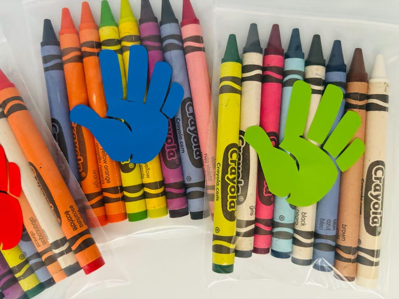 May include: Two clear plastic bags filled with Crayola crayons. The bag on the left has a blue handprint sticker and the bag on the right has a green handprint sticker. The crayons are arranged in a rainbow order.