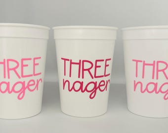 May include: Three white plastic cups with pink text that reads "THREE nager". The cups are cylindrical with a slightly flared rim. The text is in a bold, sans-serif font.