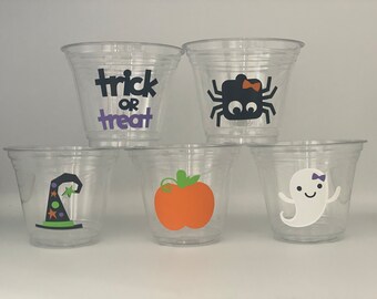 May include: Five clear plastic cups with Halloween themed designs. The designs include a witch's hat, a pumpkin, a ghost, a spider, and the words "trick or treat".