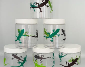 May include: Six clear glass jars with white lids, each featuring a different design of a black and green gecko. The geckos are in various poses, some with their tails curled, others with their legs outstretched.