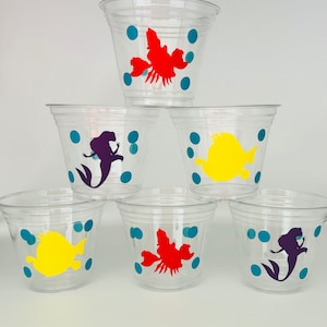 Puede incluir: Seis vasos de pl&aacute;stico transparente con siluetas de personajes coloridos. Los vasos tienen un patr&oacute;n de lunares azules. Los personajes incluyen una sirena, un pez y un cangrejo.