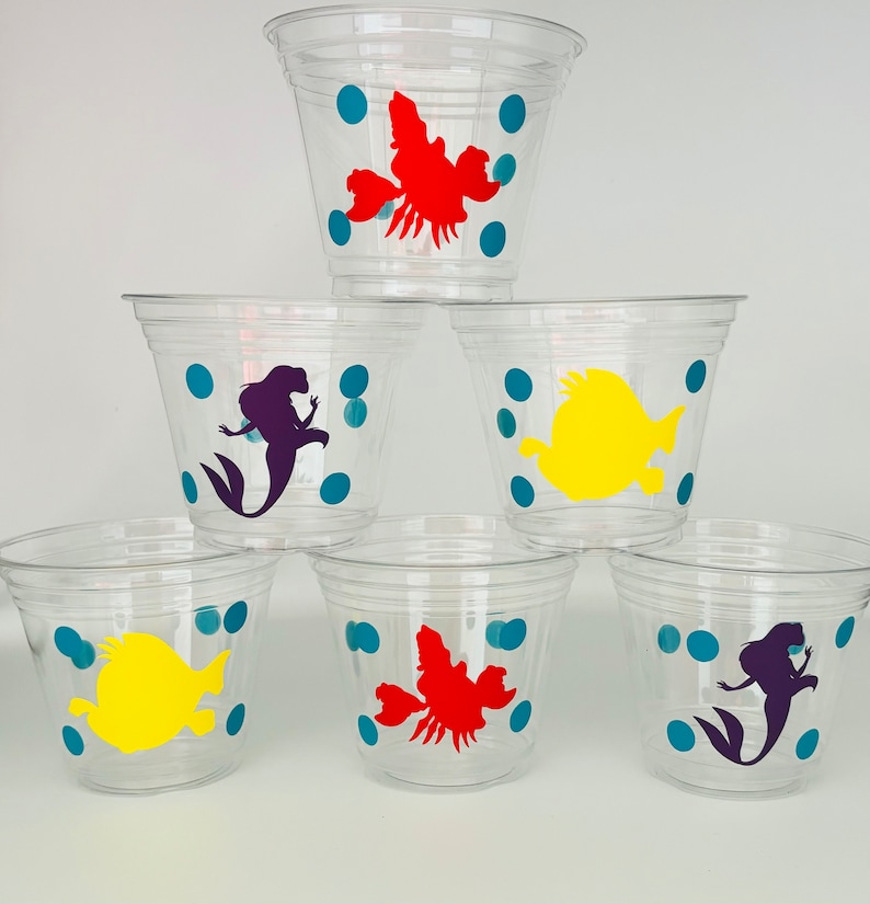 Puede incluir: Seis vasos de pl&aacute;stico transparente con siluetas de personajes coloridos. Los vasos tienen un patr&oacute;n de lunares azules. Los personajes incluyen una sirena, un pez y un cangrejo.