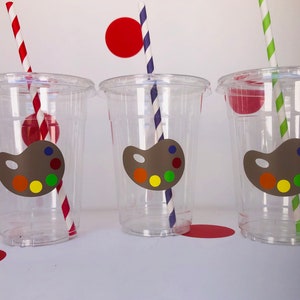 May include: Three clear plastic cups with colorful paint palette stickers and striped straws. The cups are on a white surface with red circles in the background.