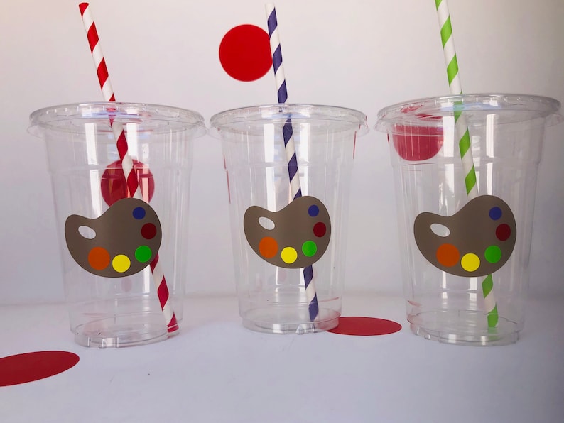 May include: Three clear plastic cups with colorful paint palette stickers and striped straws. The cups are on a white surface with red circles in the background.