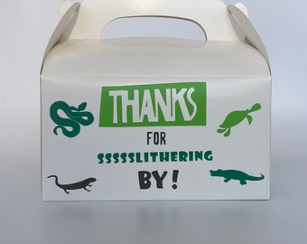 May include: White favor box with a green and black reptile theme. The box has a handle and says "Thanks for SSSSLITHERIN BY!"