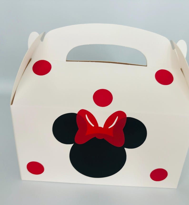Puede incluir: Caja de regalo blanca con un dise&ntilde;o de Minnie Mouse rojo y negro. La caja tiene un asa y lunares rojos.