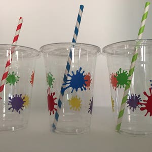 May include: Three clear plastic cups with colorful paint splatter designs and striped paper straws. The cups are perfect for a party or celebration.