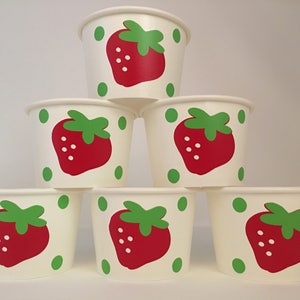 May include: Six white paper cups with a strawberry design. Each cup has a red strawberry with green leaves and white seeds. The strawberries are surrounded by green dots.