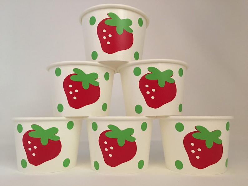 May include: Six white paper cups with a strawberry design. Each cup has a red strawberry with green leaves and white seeds. The strawberries are surrounded by green dots.