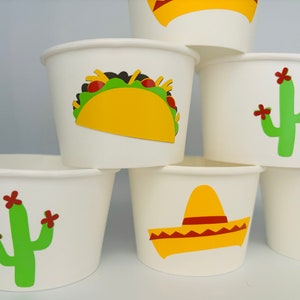 May include: White paper cups with different Mexican-themed designs. One cup has a yellow taco with green lettuce and red tomatoes. Another cup has a green cactus with red flowers. A third cup has a yellow sombrero with a red band.