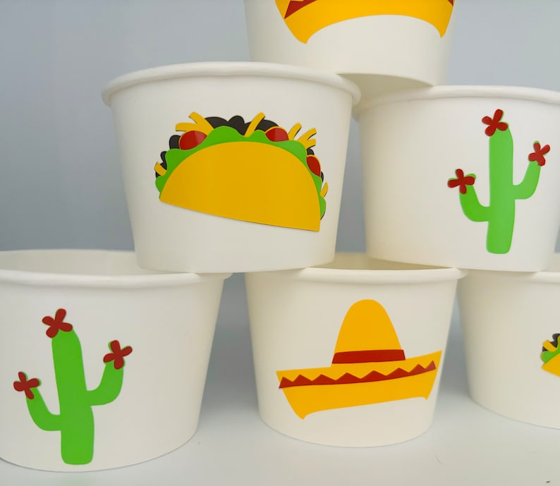 May include: White paper cups with different Mexican-themed designs. One cup has a yellow taco with green lettuce and red tomatoes. Another cup has a green cactus with red flowers. A third cup has a yellow sombrero with a red band.