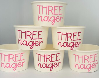 May include: A stack of six off-white paper cups with the words "THREE nager" in pink lettering. The cups are arranged in a pyramid shape, with the text clearly visible on each cup.