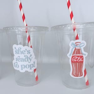 May include: Clear plastic cups with lids, each featuring a red and white striped straw. One cup has a sticker that says "she's ready to pop!" and the other has a Coca-Cola bottle sticker.