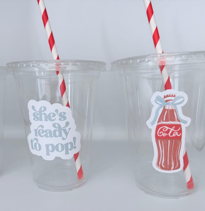 May include: Clear plastic cups with lids, each featuring a red and white striped straw. One cup has a sticker that says "she's ready to pop!" and the other has a Coca-Cola bottle sticker.