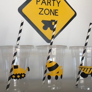 May include: Three clear plastic cups with black and white striped straws. Each cup has a yellow and black construction vehicle sticker. The sign behind the cups says "Party Zone" with a yellow and black construction truck image.