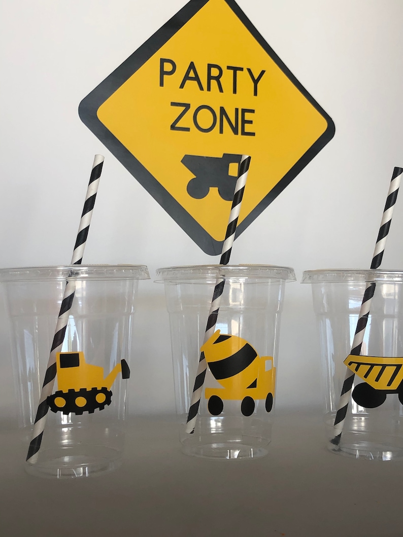 May include: Three clear plastic cups with black and white striped straws. Each cup has a yellow and black construction vehicle sticker. The sign behind the cups says "Party Zone" with a yellow and black construction truck image.