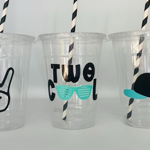 May include: Three clear plastic cups with black and white striped straws. The cups have different designs: one has a peace sign, one says "Two Cool" with sunglasses, and one has a black and blue baseball cap.