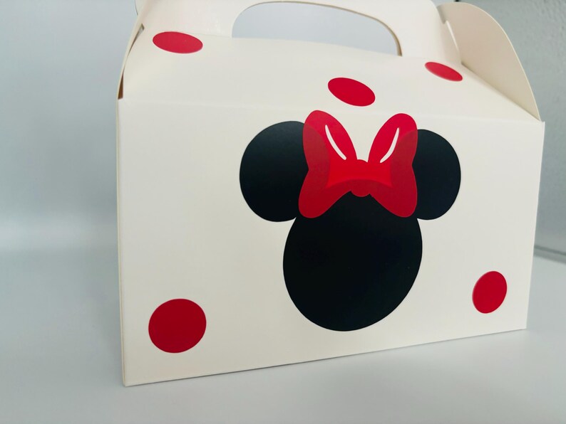Puede incluir: Caja de cart&oacute;n blanca con un dise&ntilde;o de Minnie Mouse rojo y negro. La caja tiene un asa y lunares rojos.