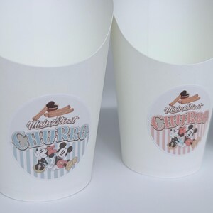 Op de afbeelding: Witte papieren voedselbakjes met een ronde sticker met de tekst Main Street CHURRO, een illustratie van churros en stripfiguurmuizen. De linkerbak heeft een blauw gestreepte achtergrond en de rechterbak heeft roze strepen.
