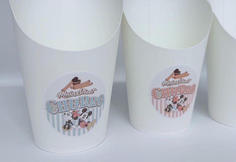 Op de afbeelding: Witte papieren voedselbakjes met een ronde sticker met de tekst Main Street CHURRO, een illustratie van churros en stripfiguurmuizen. De linkerbak heeft een blauw gestreepte achtergrond en de rechterbak heeft roze strepen.