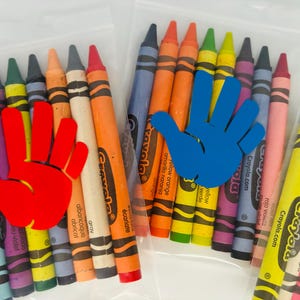 May include: A set of Crayola crayons in a clear plastic bag. The crayons are arranged in a rainbow pattern and include colors like red, orange, yellow, green, blue, purple, and black. The bag has a handprint sticker on it.
