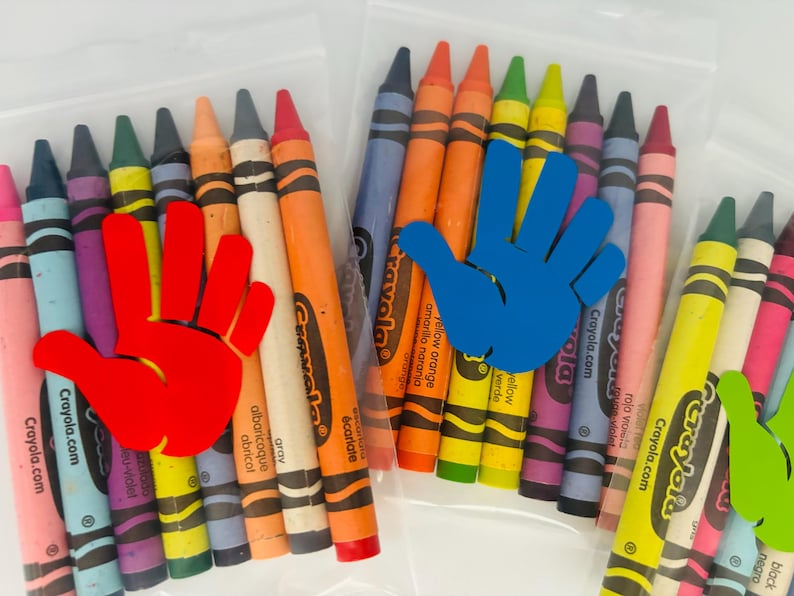 May include: A set of Crayola crayons in a clear plastic bag. The crayons are arranged in a rainbow pattern and include colors like red, orange, yellow, green, blue, purple, and black. The bag has a handprint sticker on it.