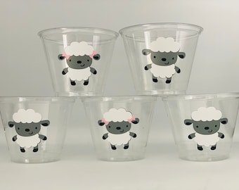 May include: Six clear plastic cups with a sheep graphic. The sheep has a white fluffy body, gray face and ears, and black feet. Some sheep have a pink bow. The cups are stacked in a pyramid shape.