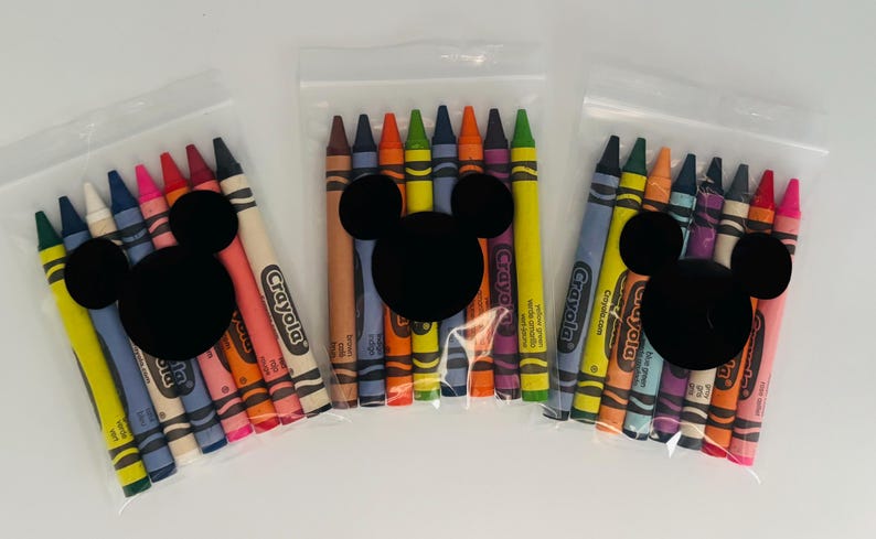 May include: Three clear plastic bags each containing a set of colorful crayons. Each bag has a black Mickey Mouse head sticker. The crayons are various colors, including yellow, blue, red, and orange. The brand name "Crayola" is visible on the crayons.